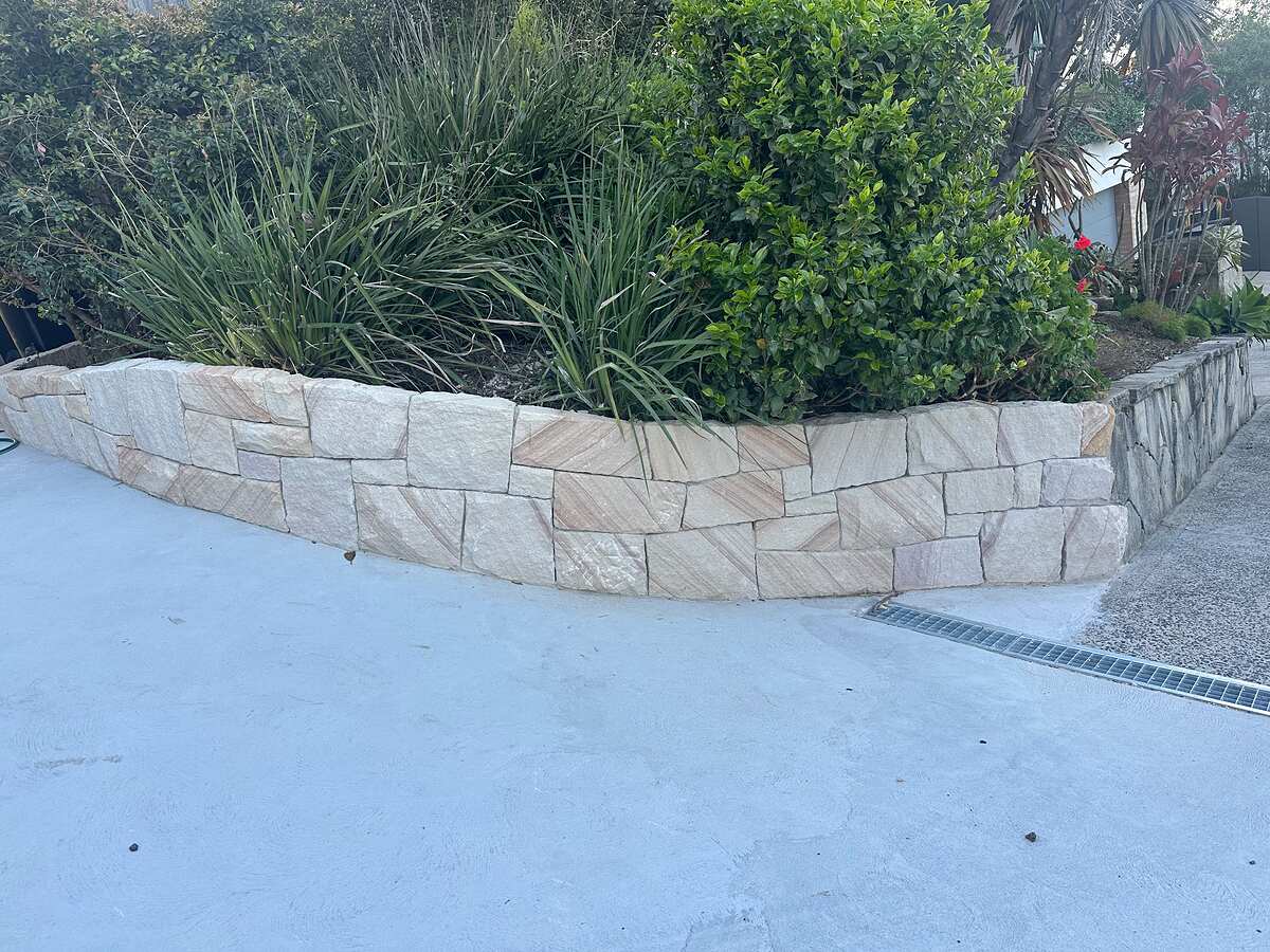 Curved sawn sandstone retaining wall holding a planted garden bed beside a concrete driveway