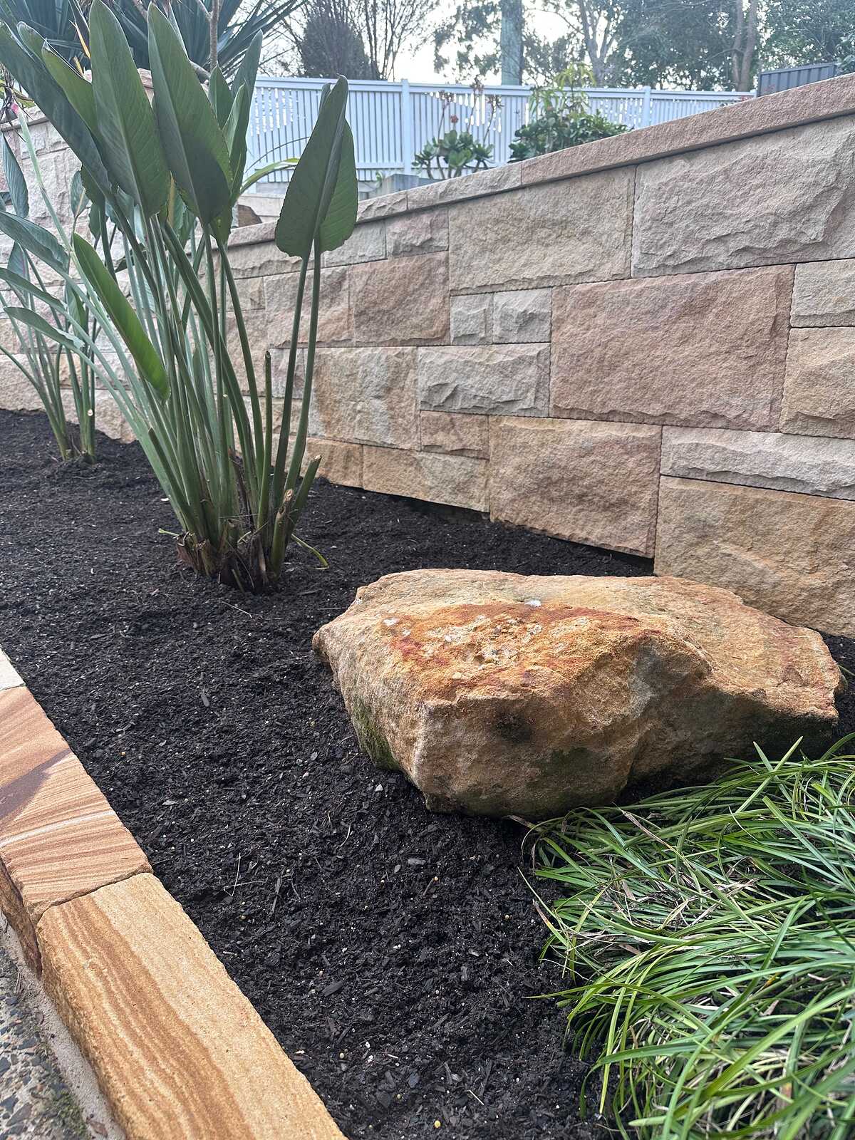 Sandstone boulder in a mulched garden bed with ashlar wall and banded sandstone edging