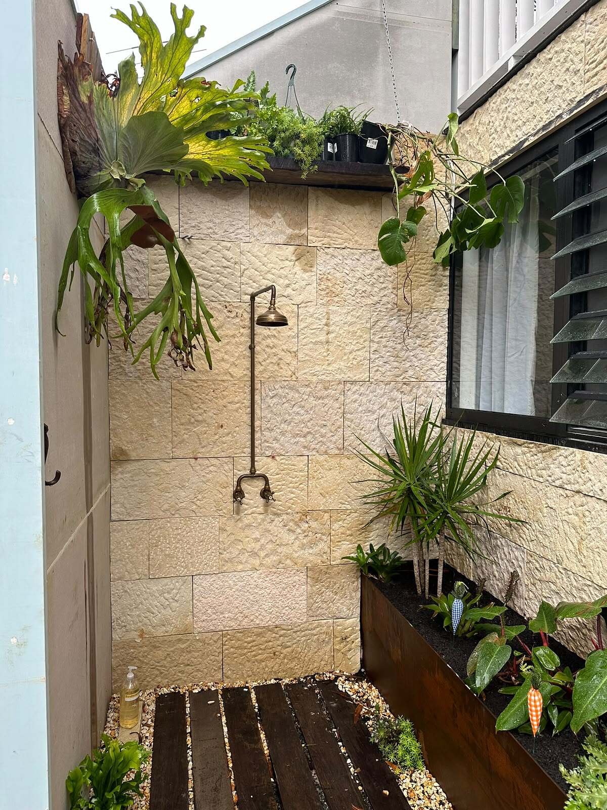 Outdoor shower with brass fittings against a split-face sandstone clad wall with ferns and planter