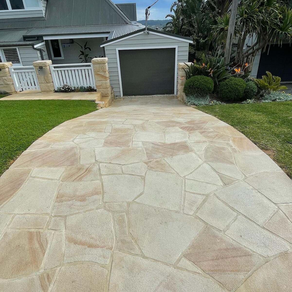 Sandstone crazy paving driveway leading to a garage with sandstone pillars and lawn