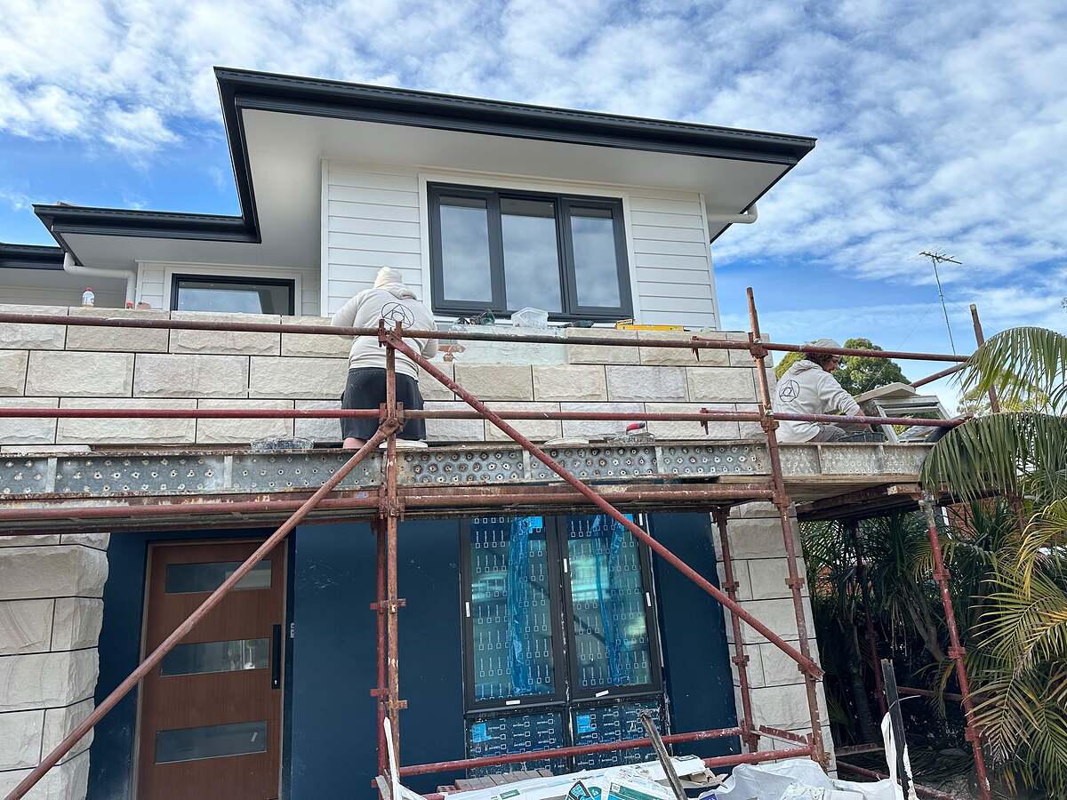 Stonemasons on scaffolding installing light split-face stone cladding on a home's upper facade